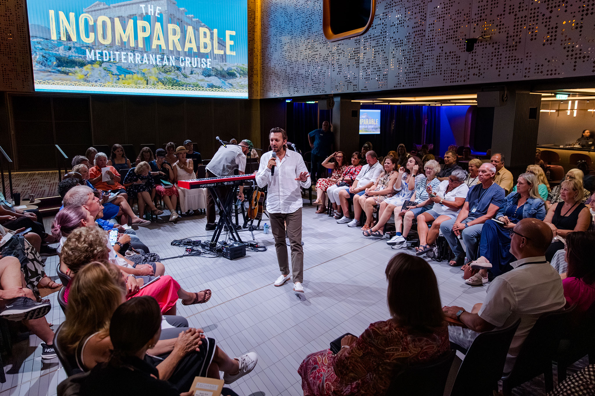 Man speaks with a microphone in the center of a seated audience during a cruise event, with instruments and event signage in the background.