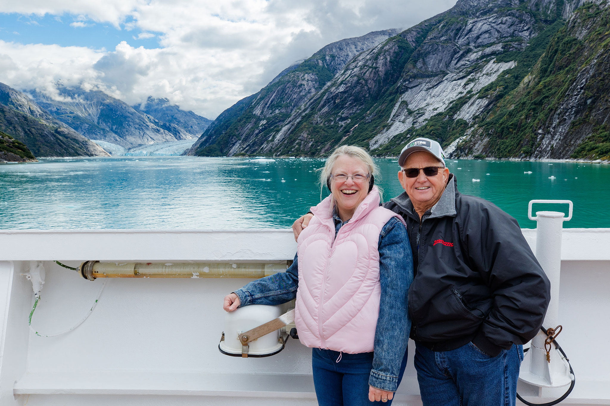 A smiling couple stands on the deck of a cruise ship with turquoise water, floating ice, and a glacier framed by steep, rocky mountains behind them in Alaska’s Glacier Bay.
