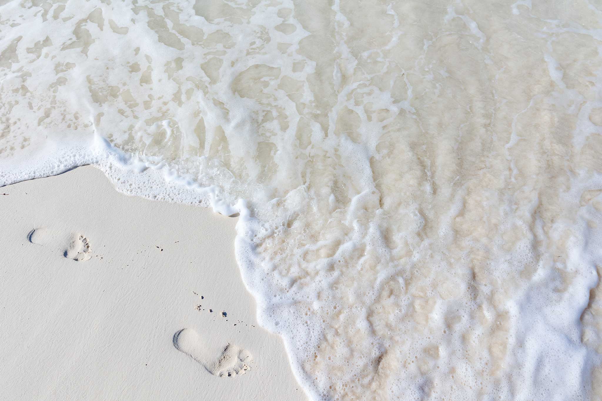 Gentle ocean waves wash over smooth white sand, partially covering a pair of footprints near the shoreline.