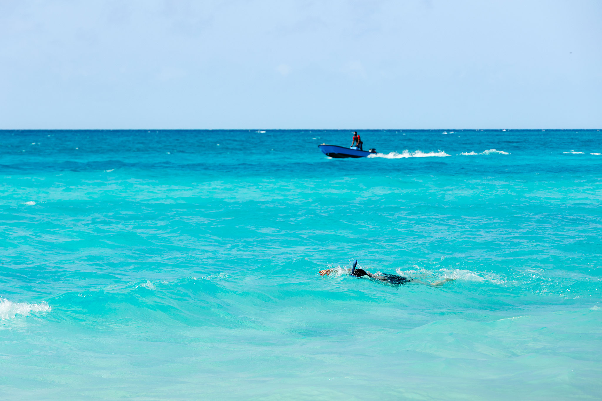 A person snorkels in bright turquoise ocean water near shore while a small motorboat cruises across the deeper blue sea in the background under a clear sky.
