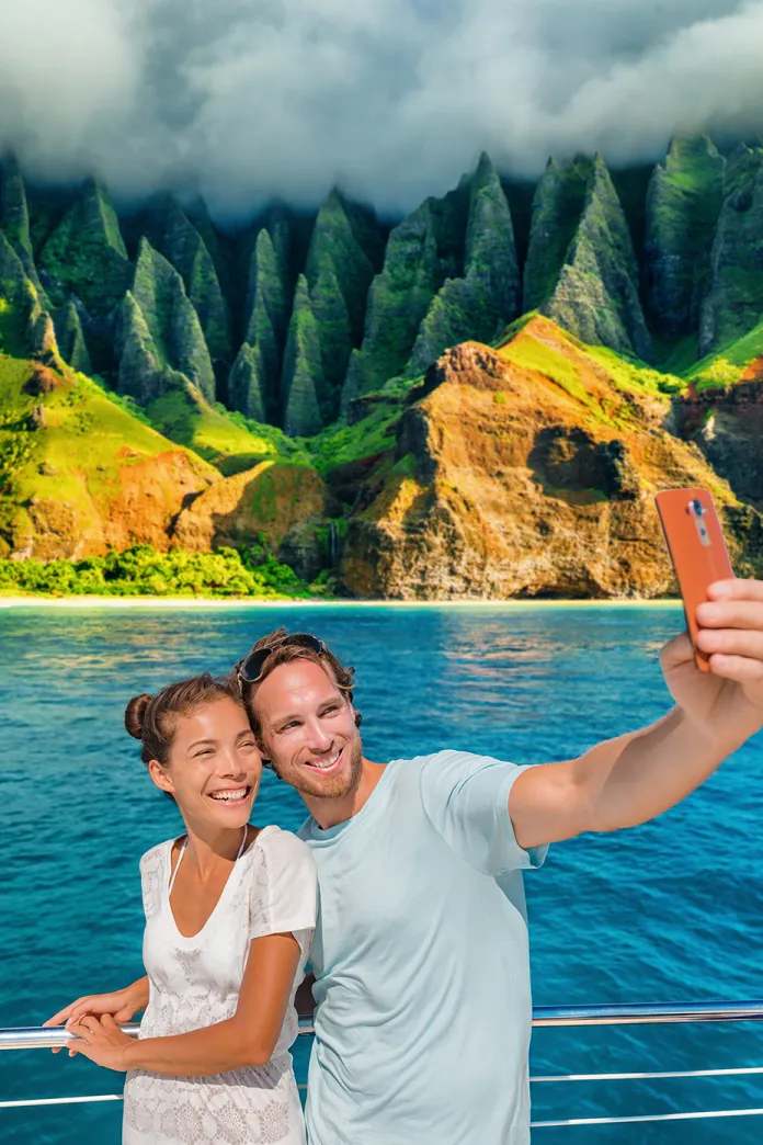 A smiling couple takes a selfie on a boat with bright blue water behind them and steep, lush green cliffs rising into low clouds in the background.